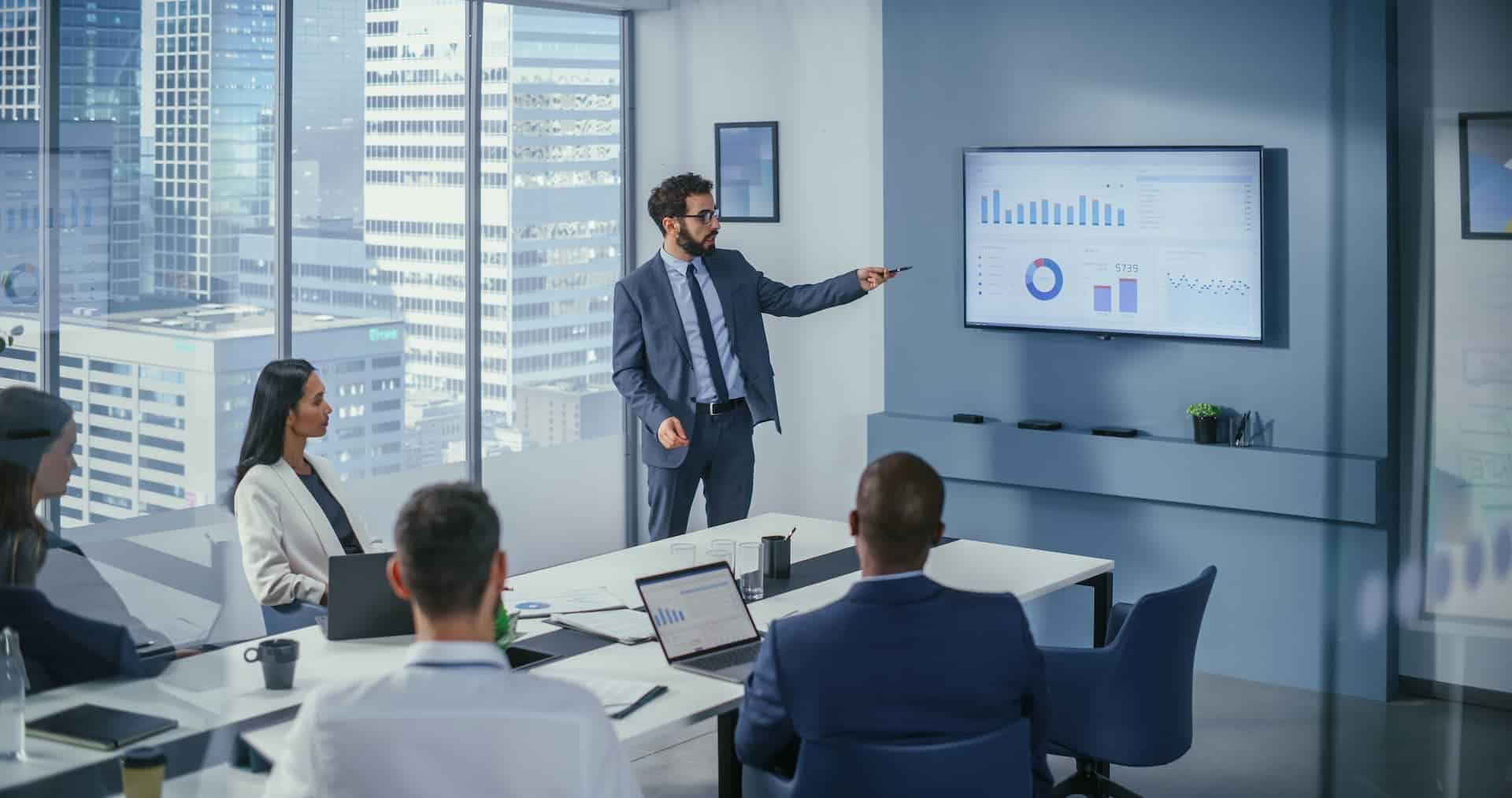 A professional meeting in a conference room where a team, seated around a table, analyzes industry trends with external orientation as a presenter highlights multiple graphs on a wall-mounted screen.
