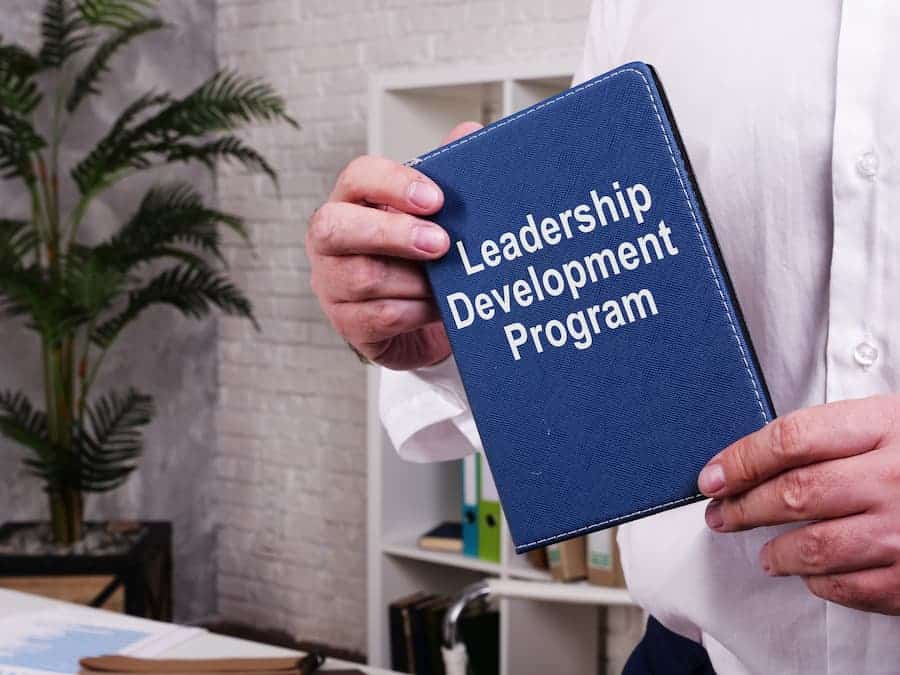 Close-up of hands holding a Leadership Development Program book in a brightly lit office with a bookcase and plant in the background.