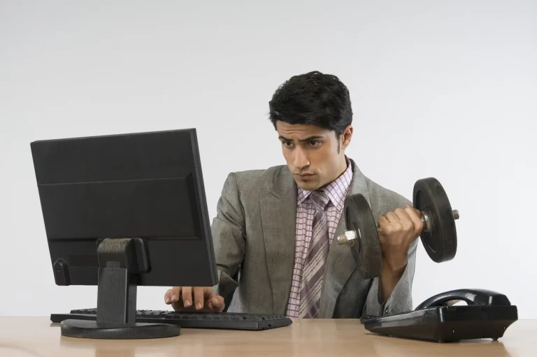 A man multitasks by lifting weights at his desk while focusing on his computer, symbolizing skill-building and productivity.