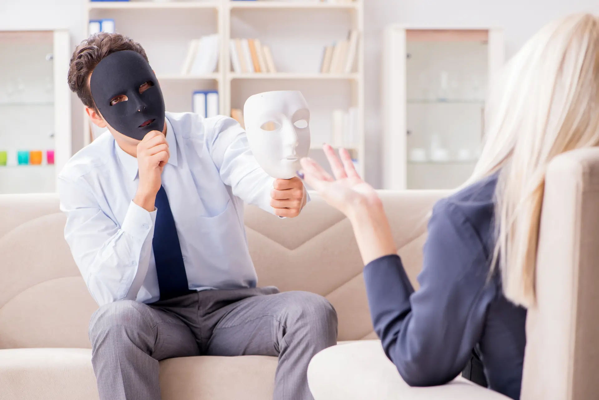 A man and woman having a conversation in a professional conversation while he wears a mask on his face, symbolizing inauthenticity or self-protection.