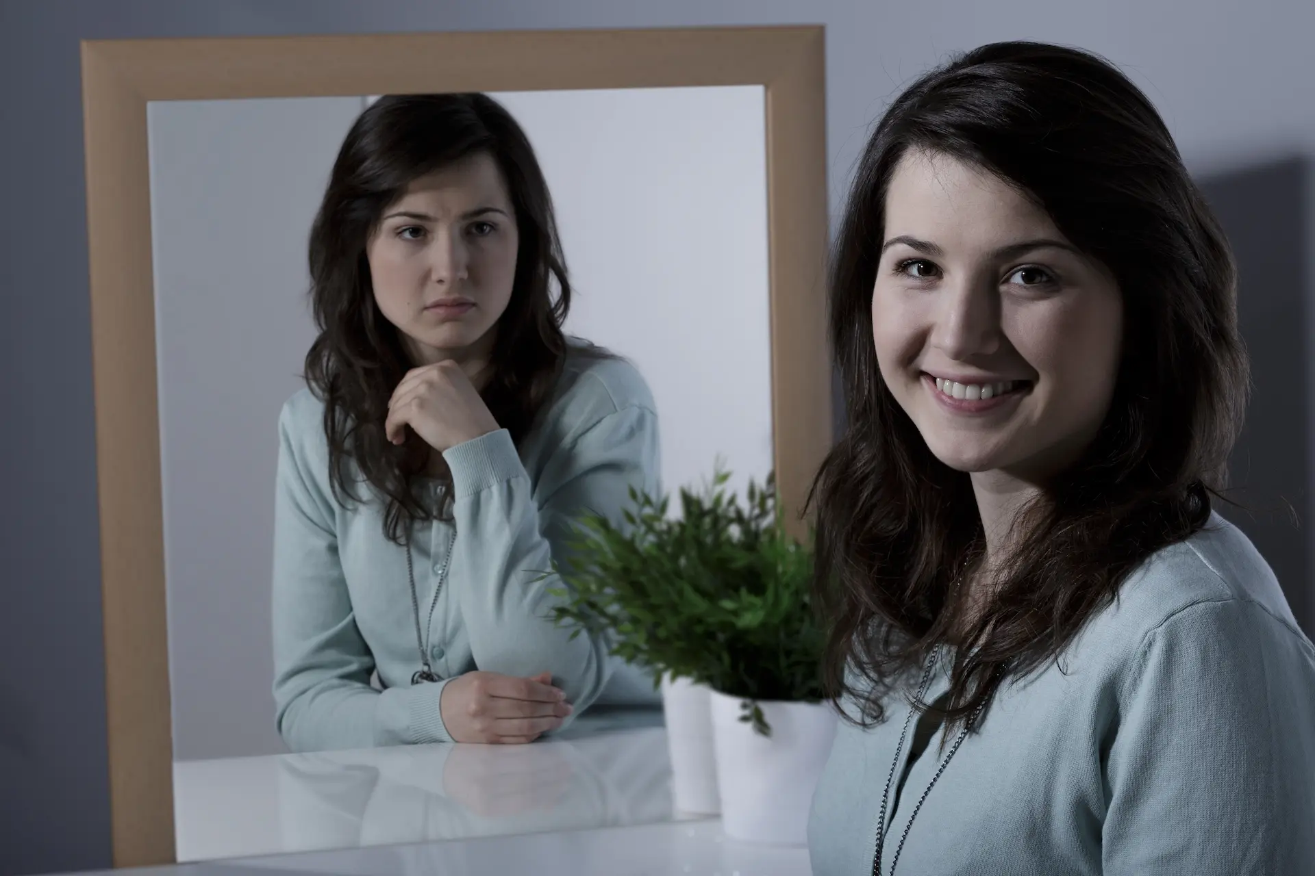 A professional woman smiling towards the camera with her reflection in the mirror showing anger, symbolizing the masks we wear in the workplace.