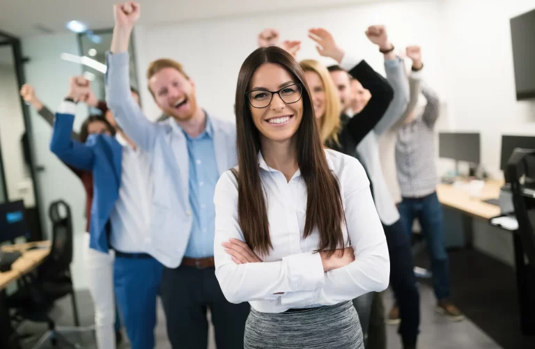 A woman with her arms crossed and smiling while her team on the back cheer her on, showcasing the importance of developing a network of support for professional success.