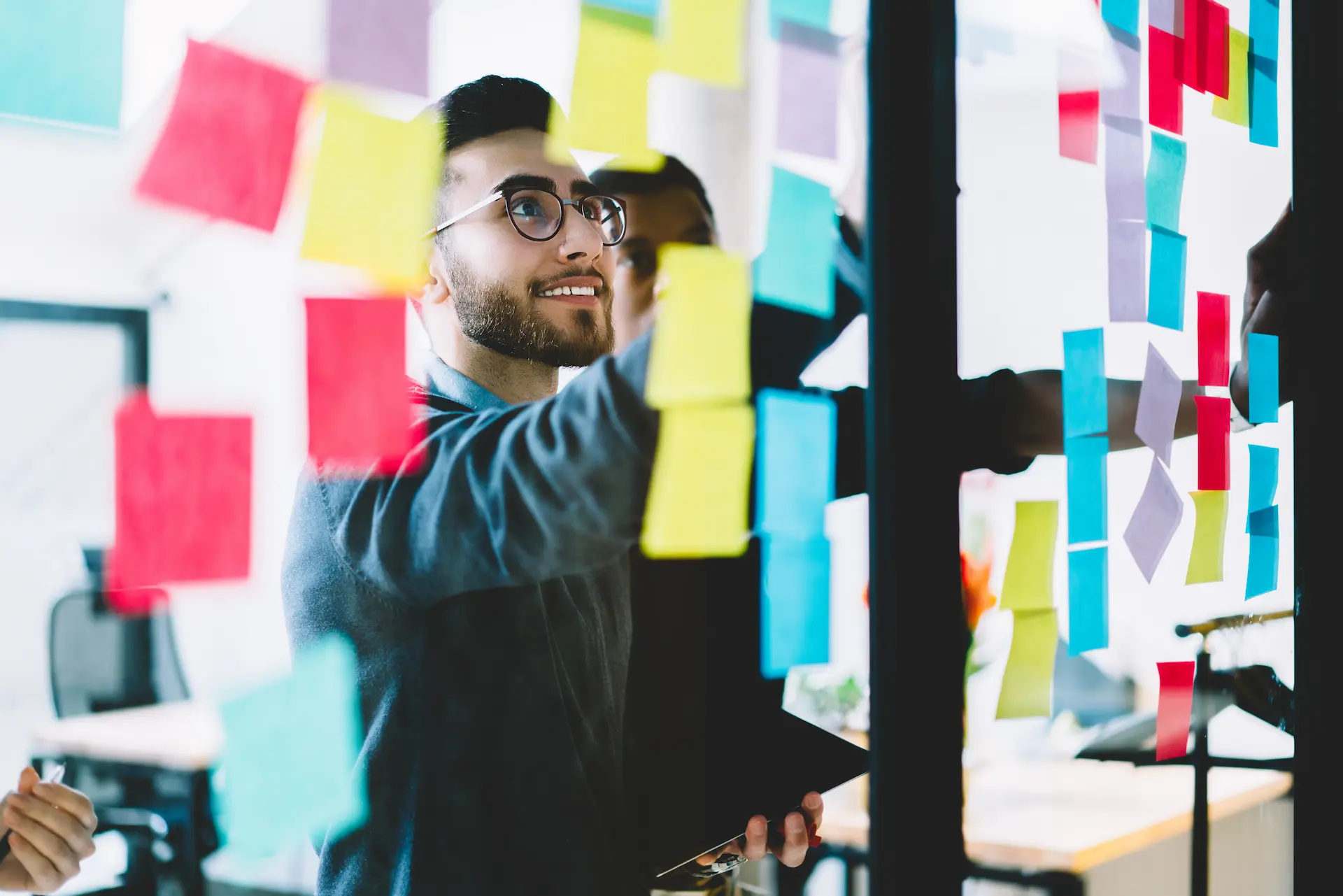 A man and his team brainstorming and planning, writing ideas on colorful Post-it notes affixed to a glass window, illustrating the collaborative stage of project execution.