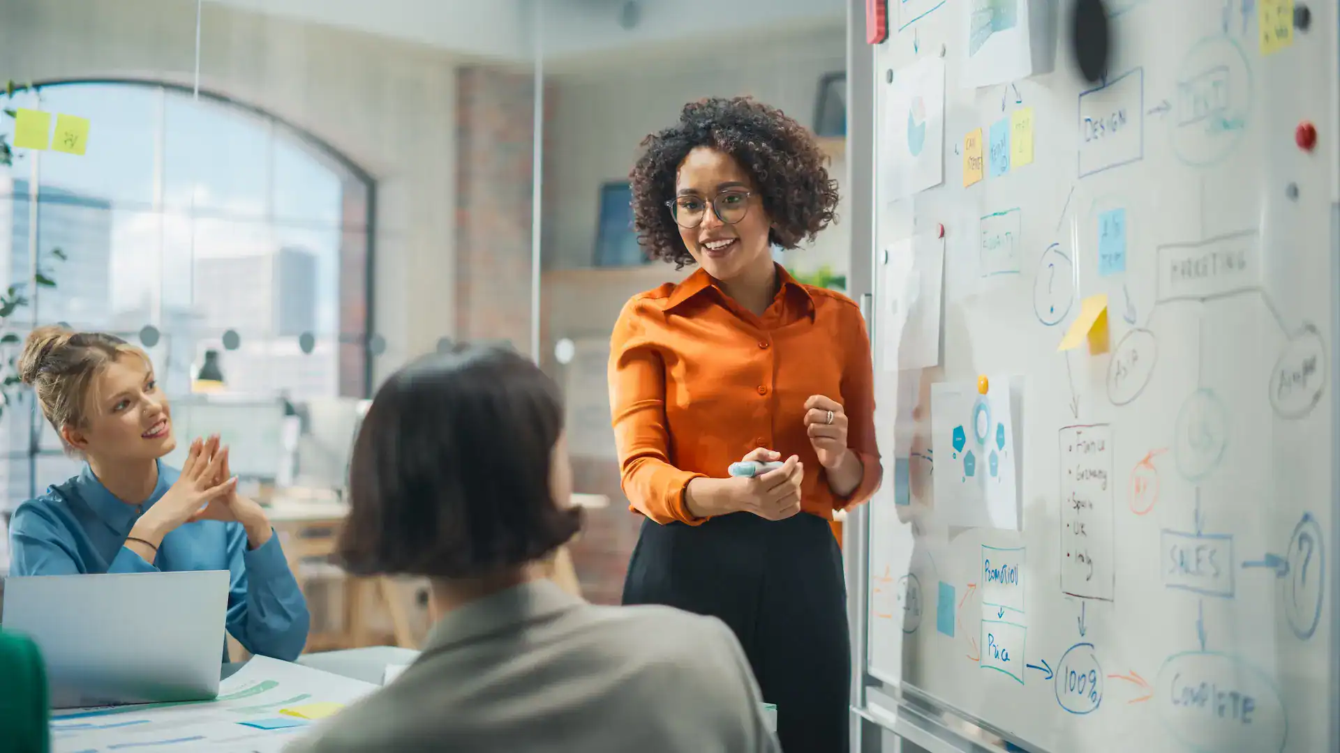 A woman leads a whiteboard brainstorming session, collaborating with a team to build strategic capabilities.
