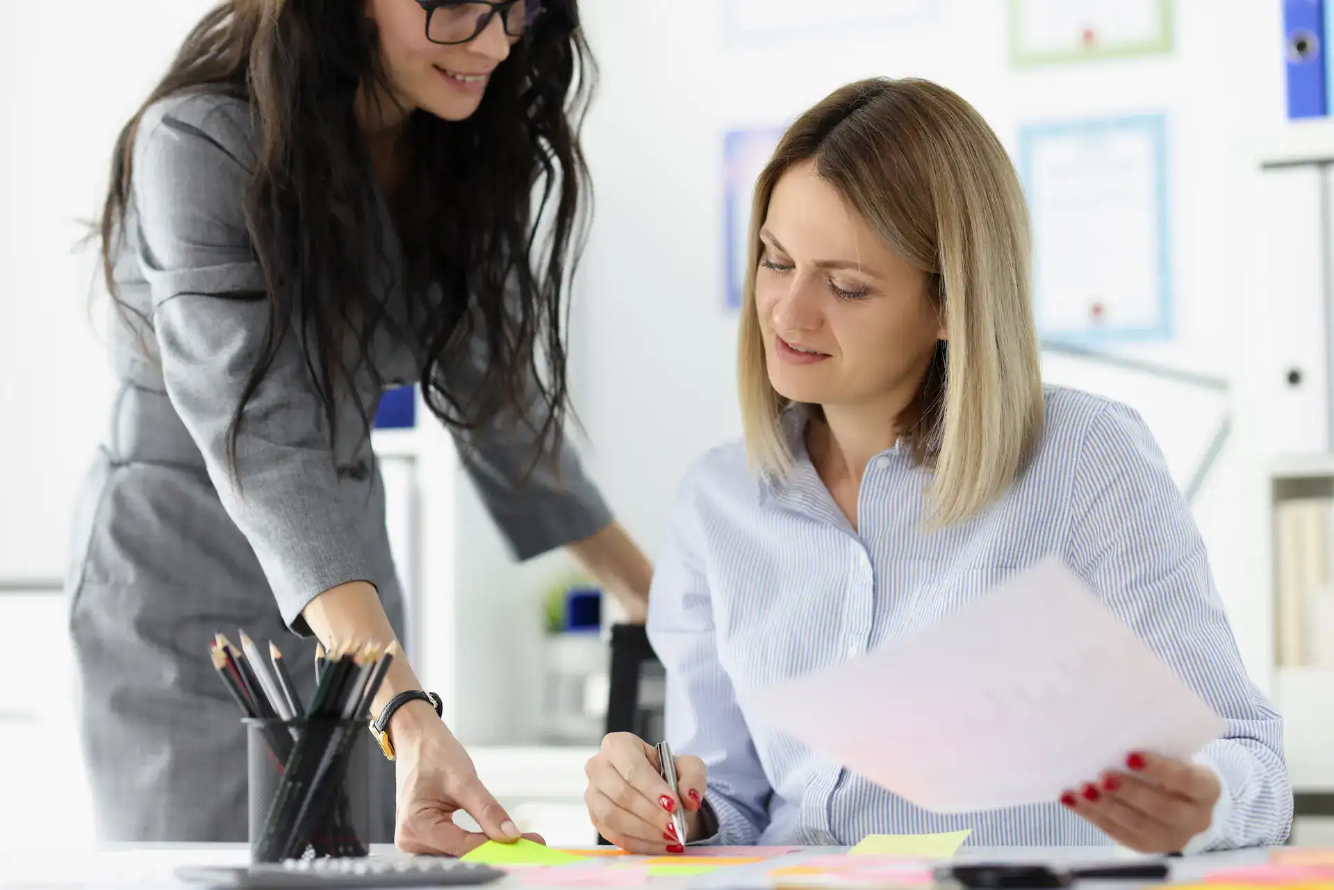 A woman passes a Post-it to a coworker, symbolizing a casual act of delegation in a team setting.