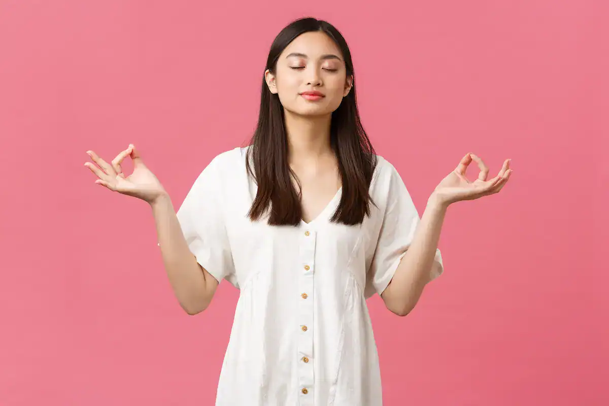Calm young woman meditating with eyes closed and hands in a relaxed mudra pose, symbolizing mindfulness and inner balance, on a pink background.