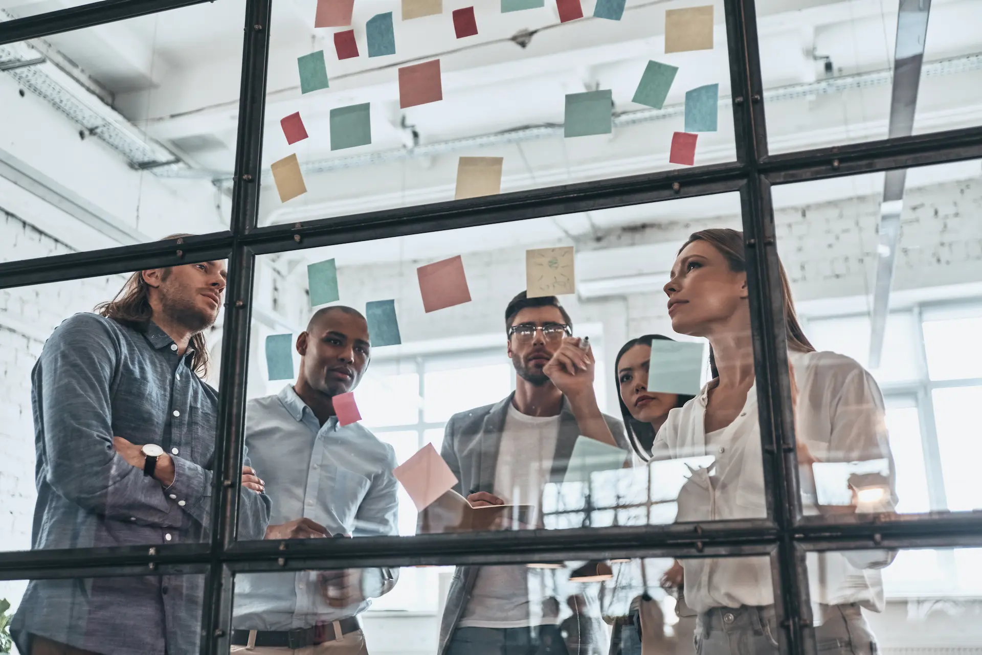 A group of coworkers using markers on a glass wall covered in sticky notes, applying lean principles to brainstorm ideas and reduce process waste.