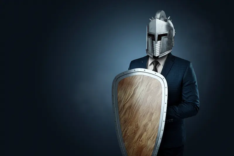 Business professional wearing a metal helmet and holding a wooden shield, symbolizing emotional masks and self-protection in the workplace.