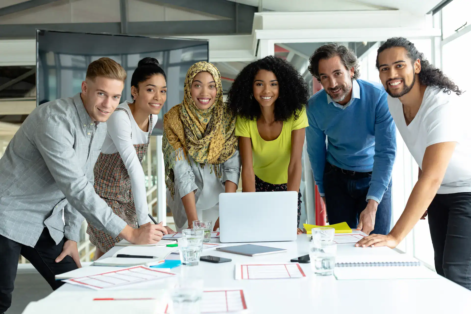 A diverse group of professionals gathered around a table, smiling and collaborating with a laptop and documents in front of them, representing effective leadership through teamwork and a mix of skills.
