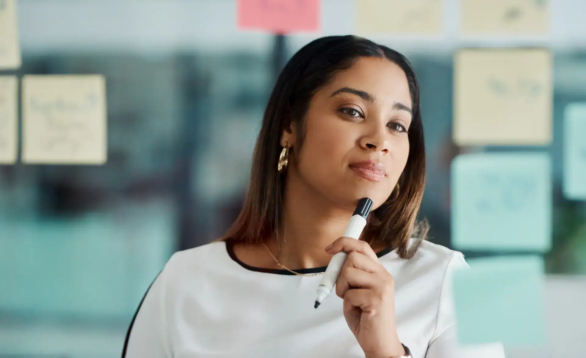 A thoughtful woman holding a marker while looking at sticky notes on a glass wall, representing reflection, planning, and the strategic thinking behind building a diverse skill set.