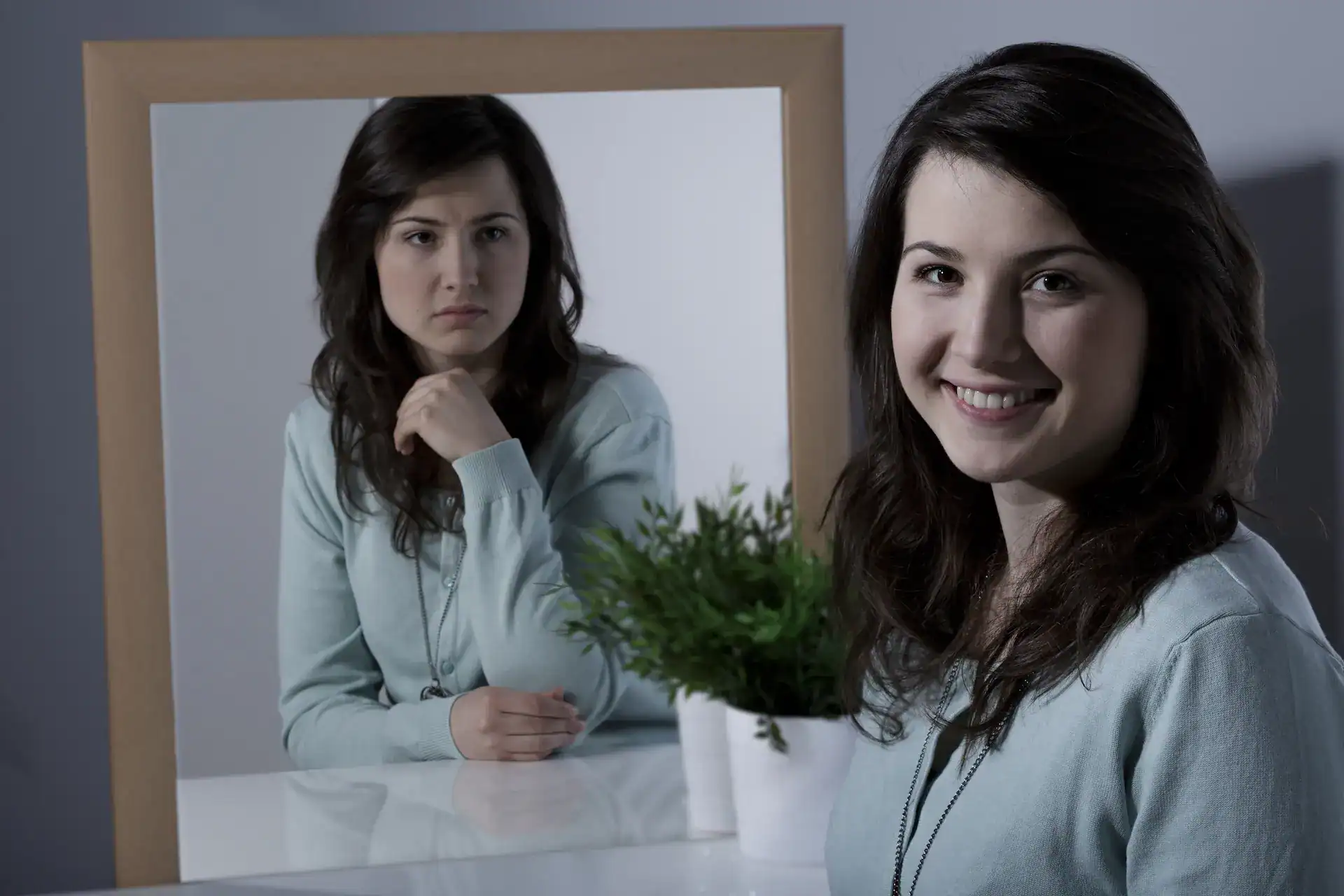 A woman smiling to the camara while her reflection in a mirror shows her upset at herself, showing how our inner critic can be judgmental of ourselves.