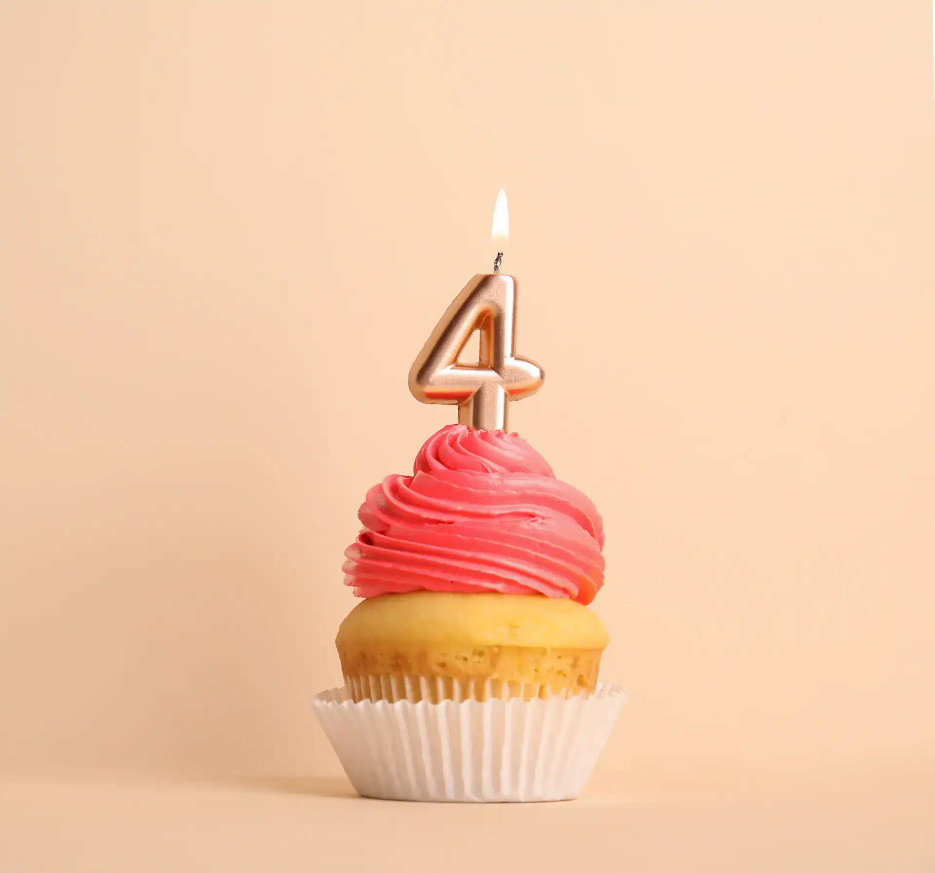 Vanilla cupcake with pink frosting and a lit number four candle, set against a beige background.