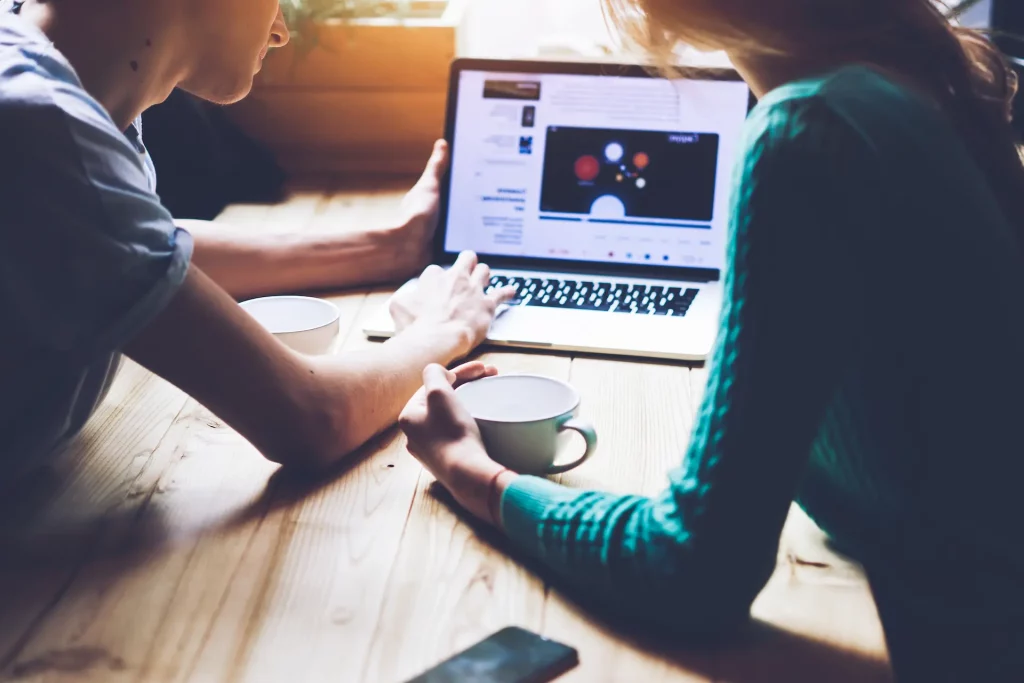 Two people sitting over a laptop as one helps the other build a strong LinkedIn profile.