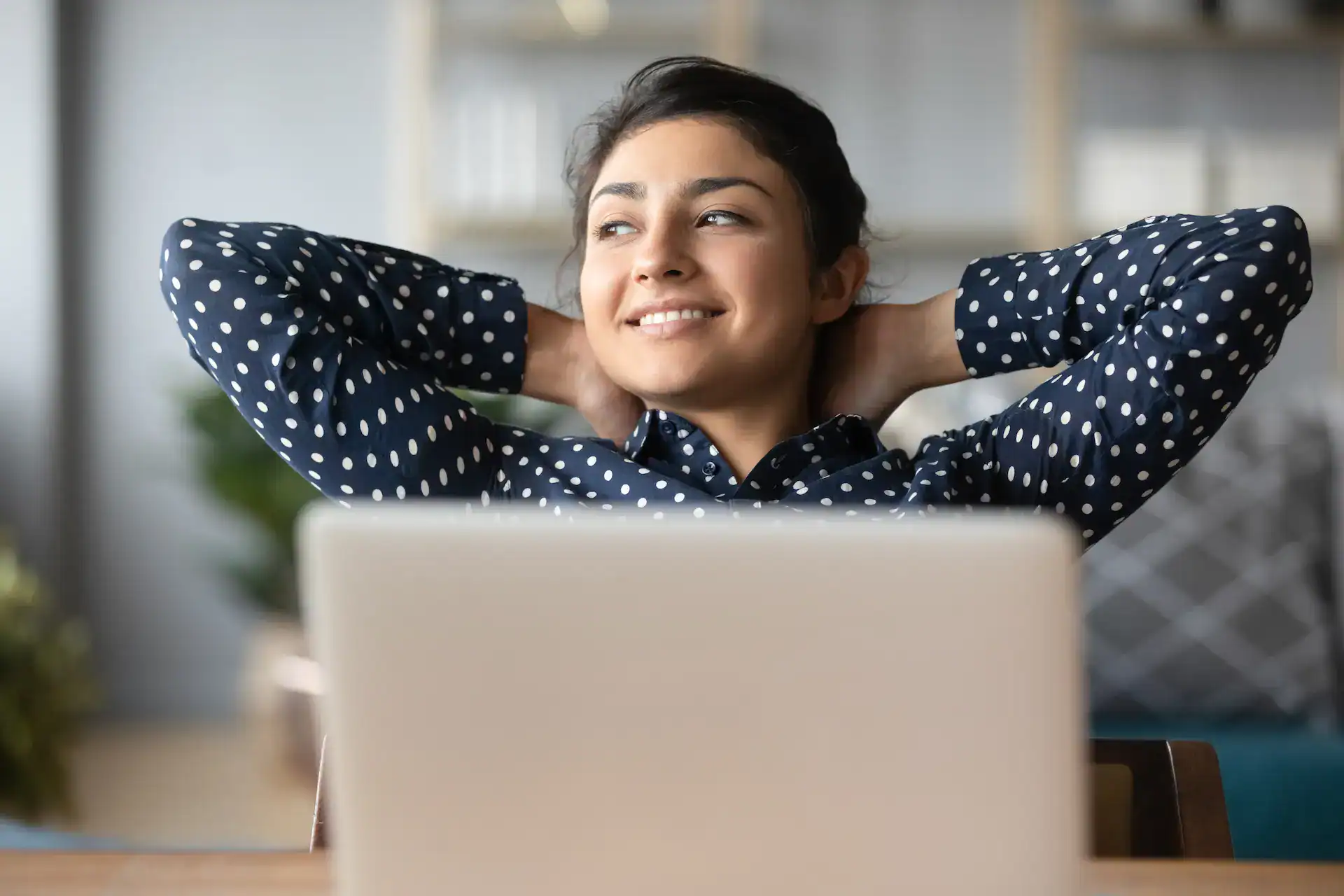 A woman leans back in her chair with her hands behind her head, smiling contentedly while looking away from her laptop, conveying a sense of achievement and relaxation after successful goal setting.