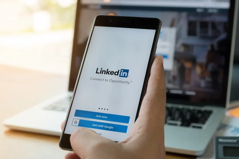 A hand holding a phone over a laptop, both displaying the LinkedIn logo on their screens.