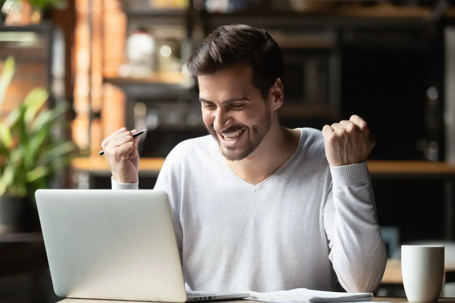 A man excitedly raises his fists in front of his laptop, celebrating a successful outcome, which could be the result of effective goal setting.