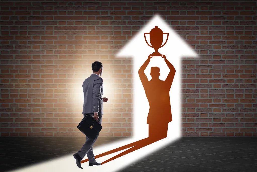 A man walks towards an illuminated shadow on a wall in the shape of an upward-pointing arrow that shows him holding up a trophy, representing the pursuit of reward and personal recognition.