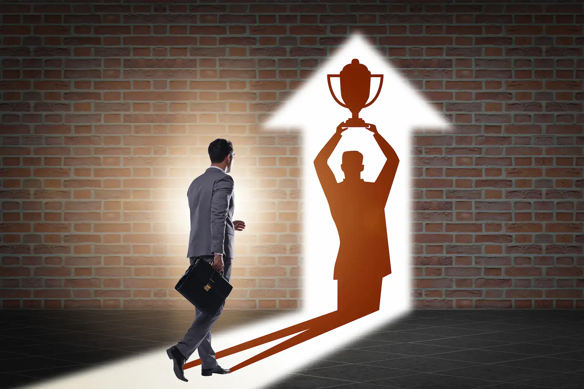 A man walks towards an illuminated shadow on a wall in the shape of an upward-pointing arrow that shows him holding up a trophy, representing the pursuit of reward and personal recognition.