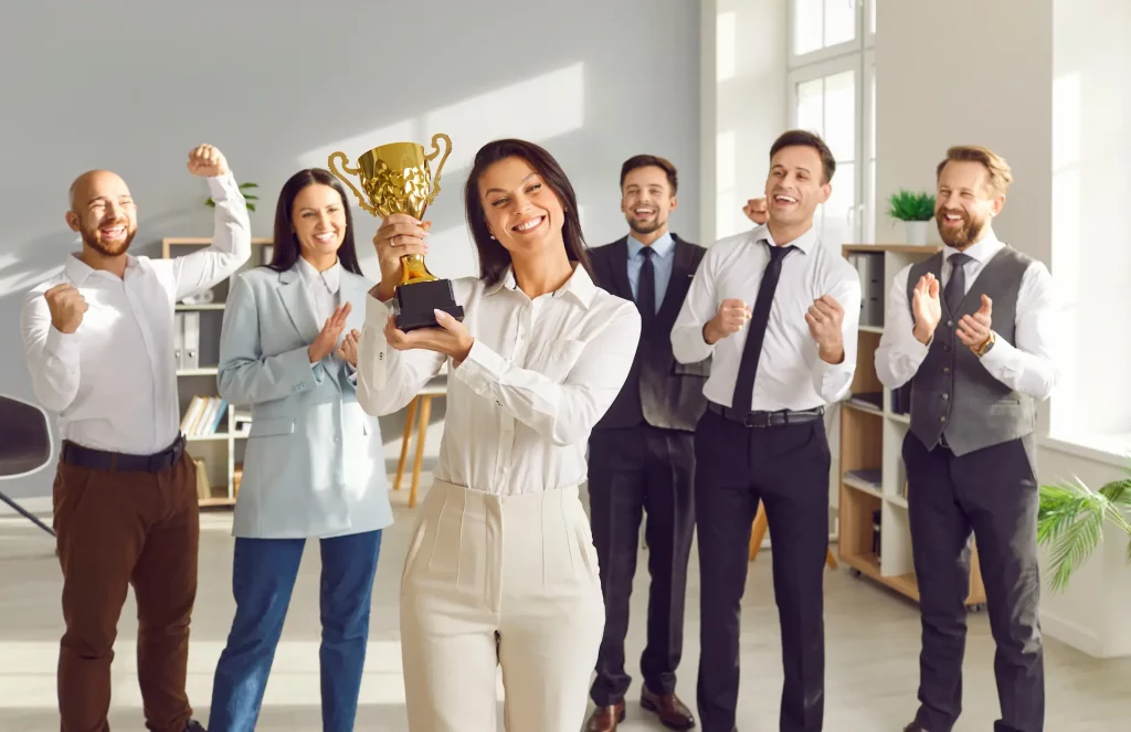 A diverse group of smiling people celebrate with one woman holding a trophy, signifying collective achievement and individual recognition.