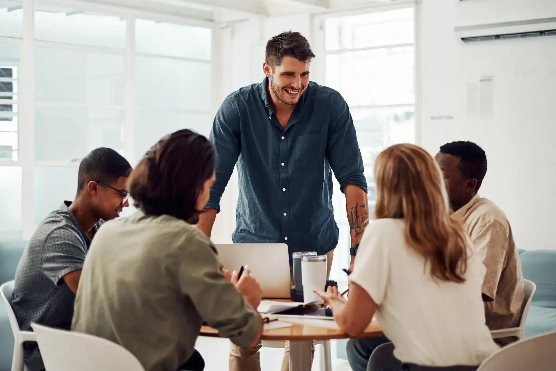 A male team leader smiles and stands behind a desk, leading a diverse group of employees who understand their role clarity.