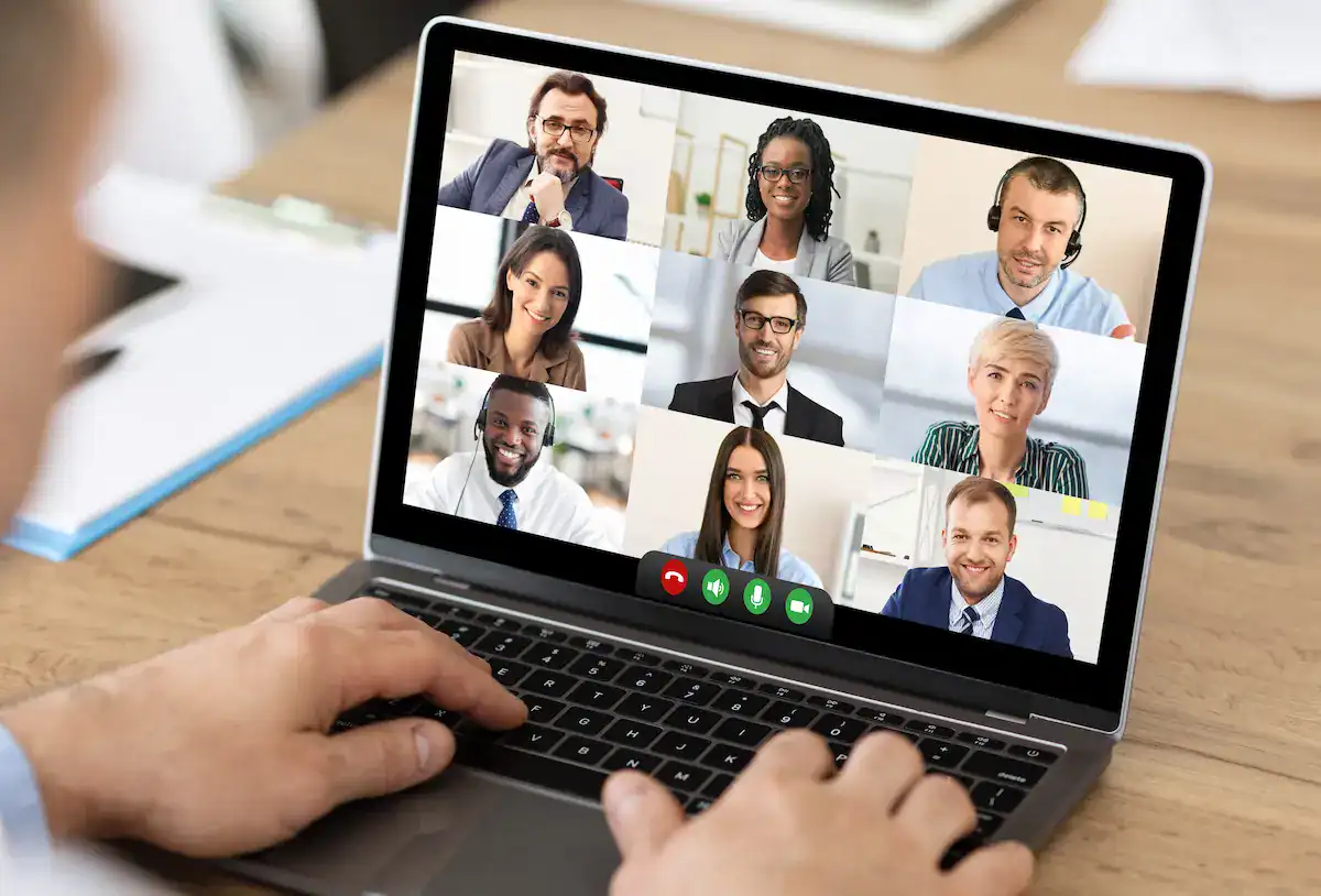 A person's hands are visible on a keyboard as a laptop screen displays a grid of nine individuals in a video conference, illustrating remote presence in leadership.