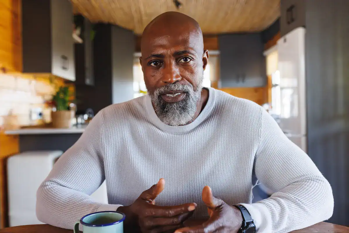 A mature man with a beard looks directly at the camera, gesturing with his hands, creating a sense of remote presence.