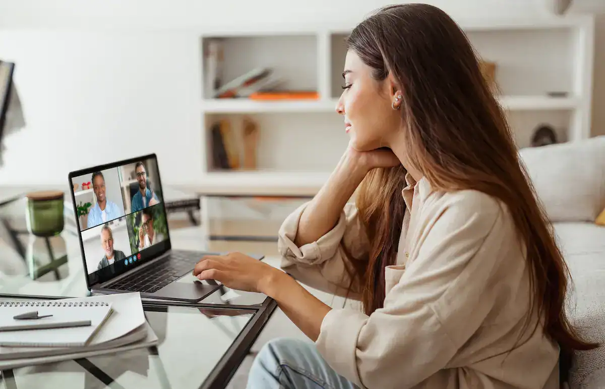 A woman sits at a desk with her laptop, participating in a video call with four other participants, showcasing her technical home setup for remote work and presence.