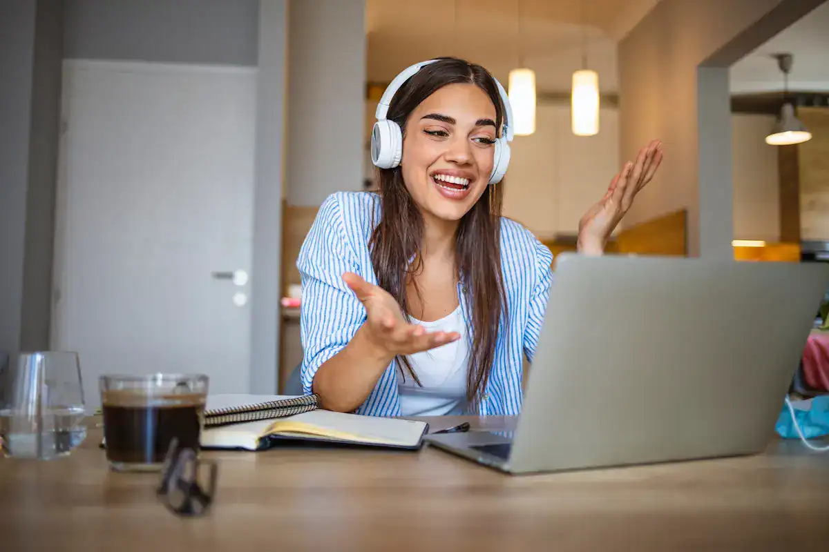A woman wearing headphones smiles and gestures at her laptop screen during a video call, capturing the essence of remote presence.