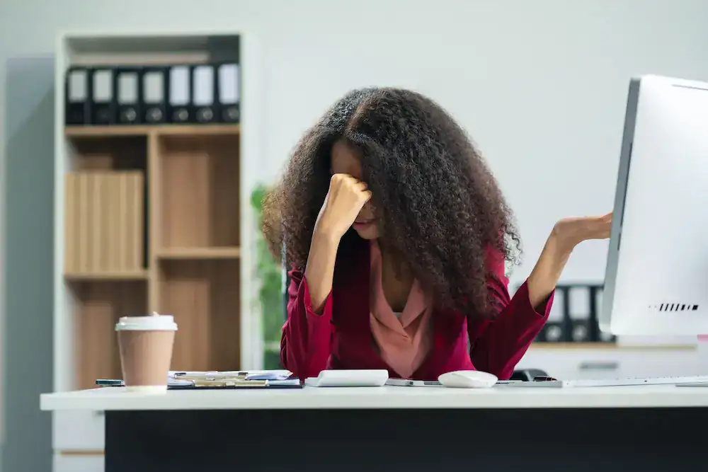 A woman sits at her desk, looking frustrated and holding her hand to her head, representing how setbacks can feel overwhelming.