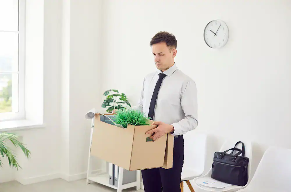 A man carries a box of belongings, showing a person leaving a job after experiencing setbacks.