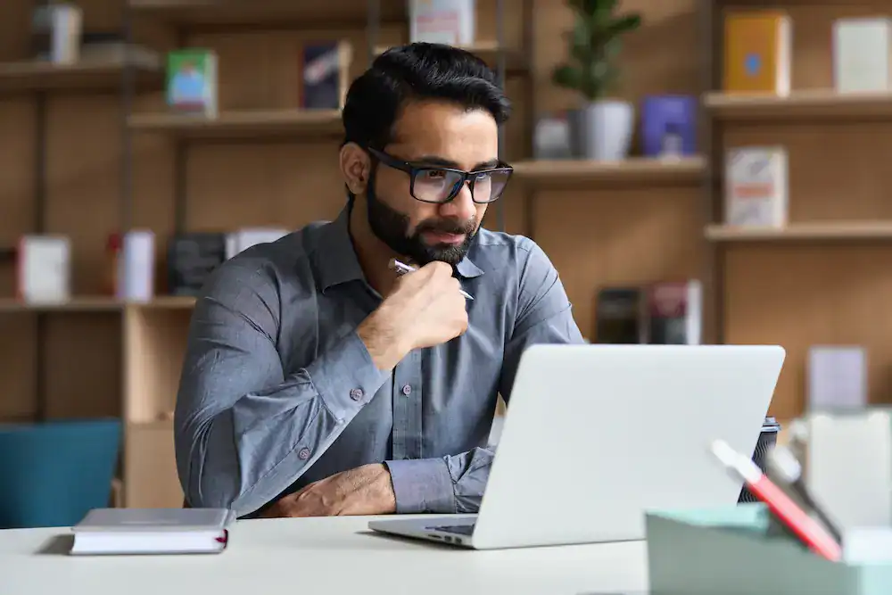 A man looking thoughtfully at his laptop with a pen in hand shows a moment of concentration crucial for mental fitness.