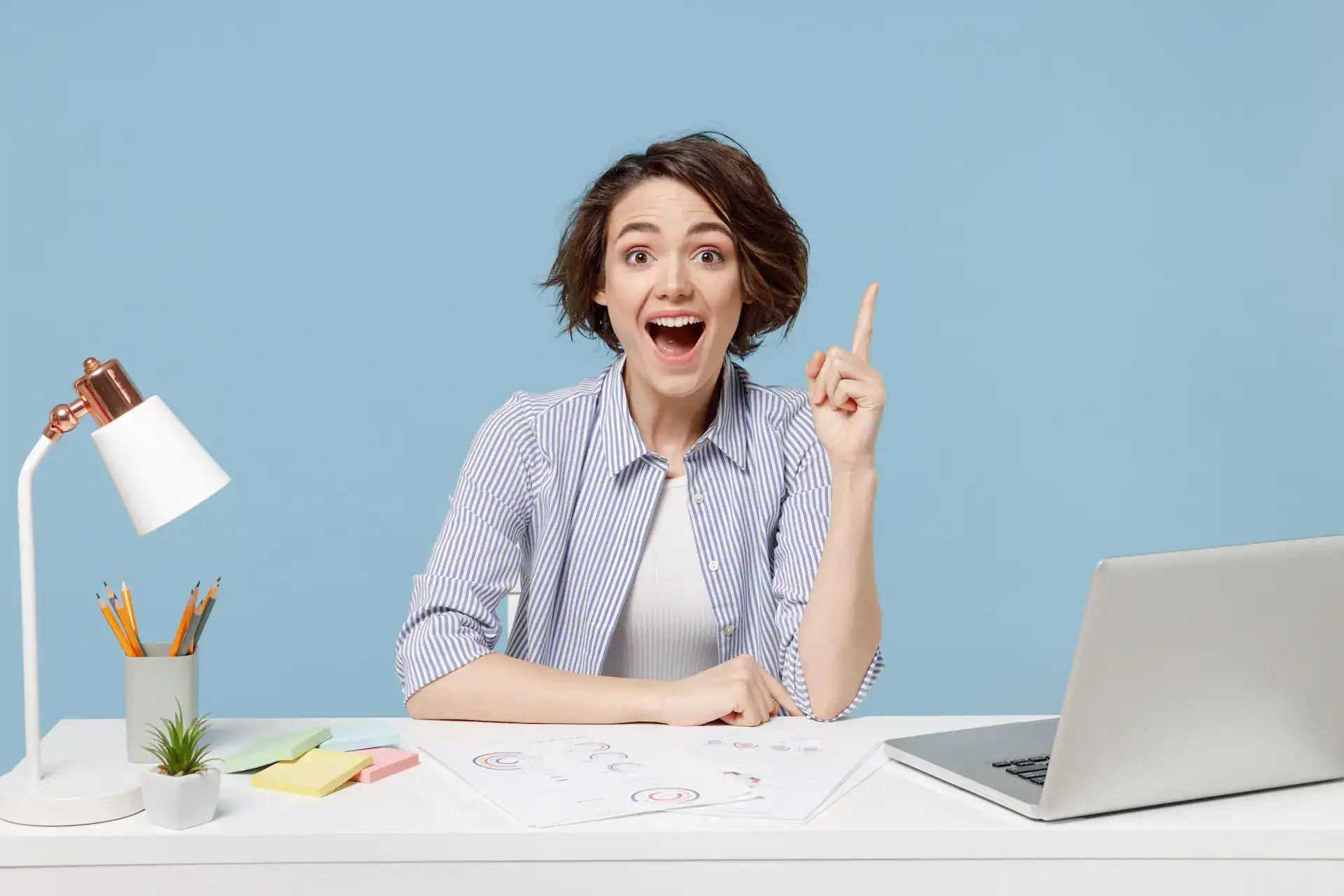 Smiling young professional at a desk raises her index finger as an idea strikes, with a laptop, charts, pencils, and a desk lamp on a blue background.
