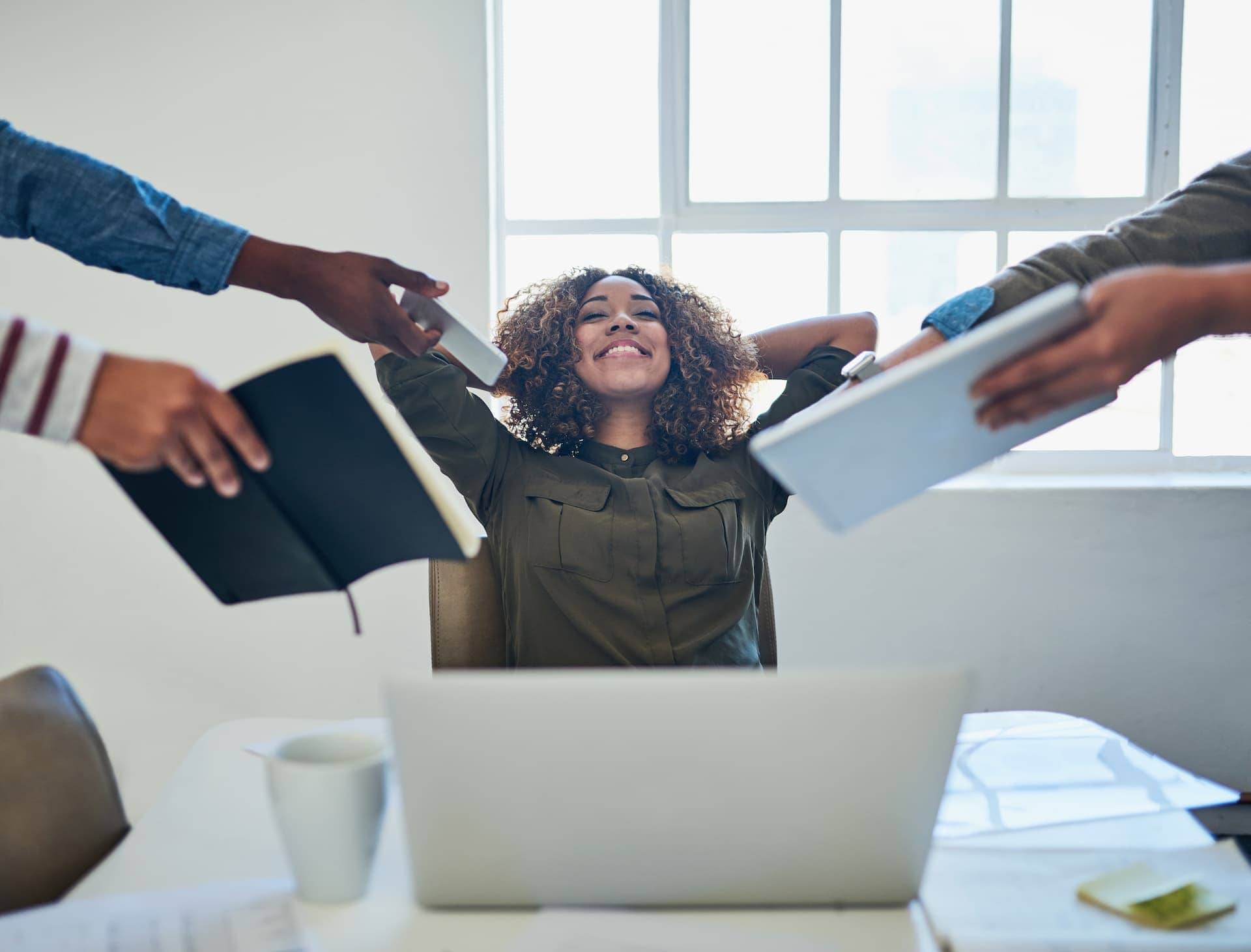 A woman calmly stretching with her arms behind her head at her desk, staying relaxed despite being handed multiple tasks.