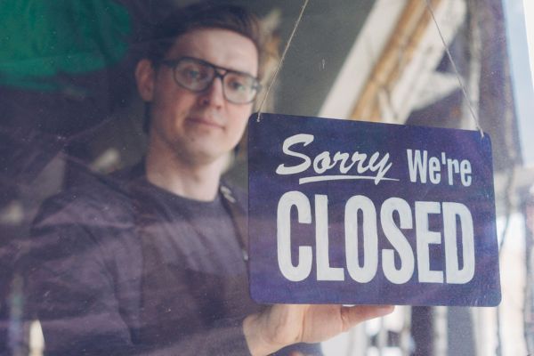 Man in glasses holding a Sorry We're Closed sign in a shop window.