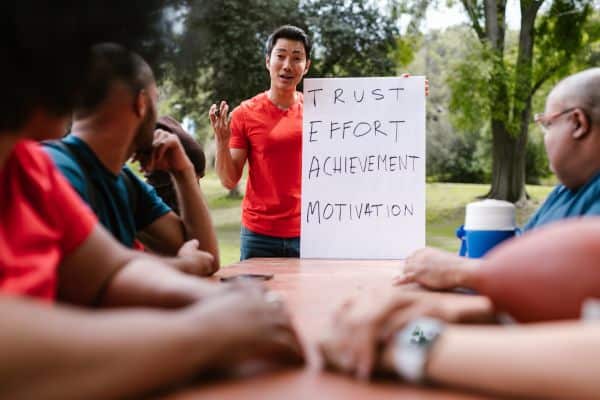 Leader in front of a whiteboard listing trust, effort, achievement and motivation, talking to a team at an outdoor table.