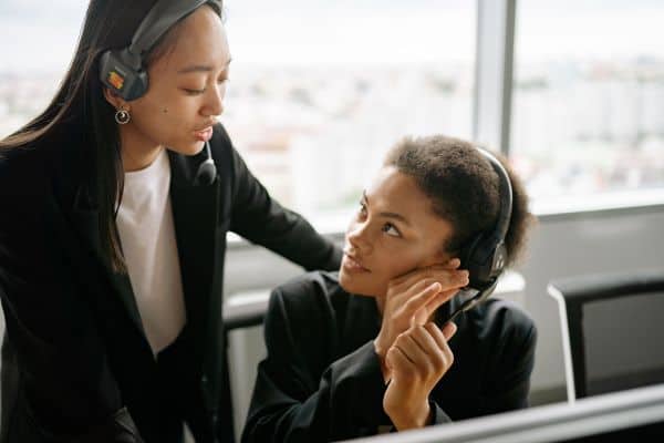 Colleague wearing a headset leaning in to support a coworker at her desk.